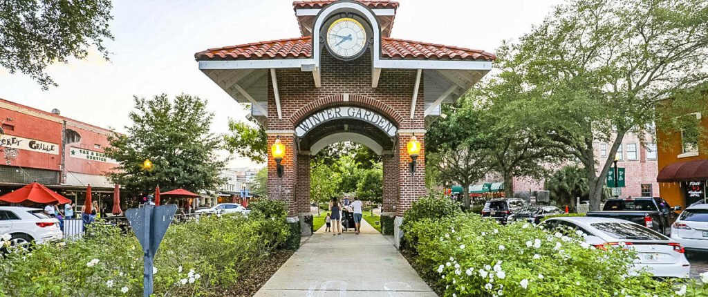 A large clock on a brick archway reading "WINTER GARDEN" over a paved path through hedges with white flowers on them in Winter Garden, Florida. Winter Garden, Florida is a location served by Johannessen Lights.